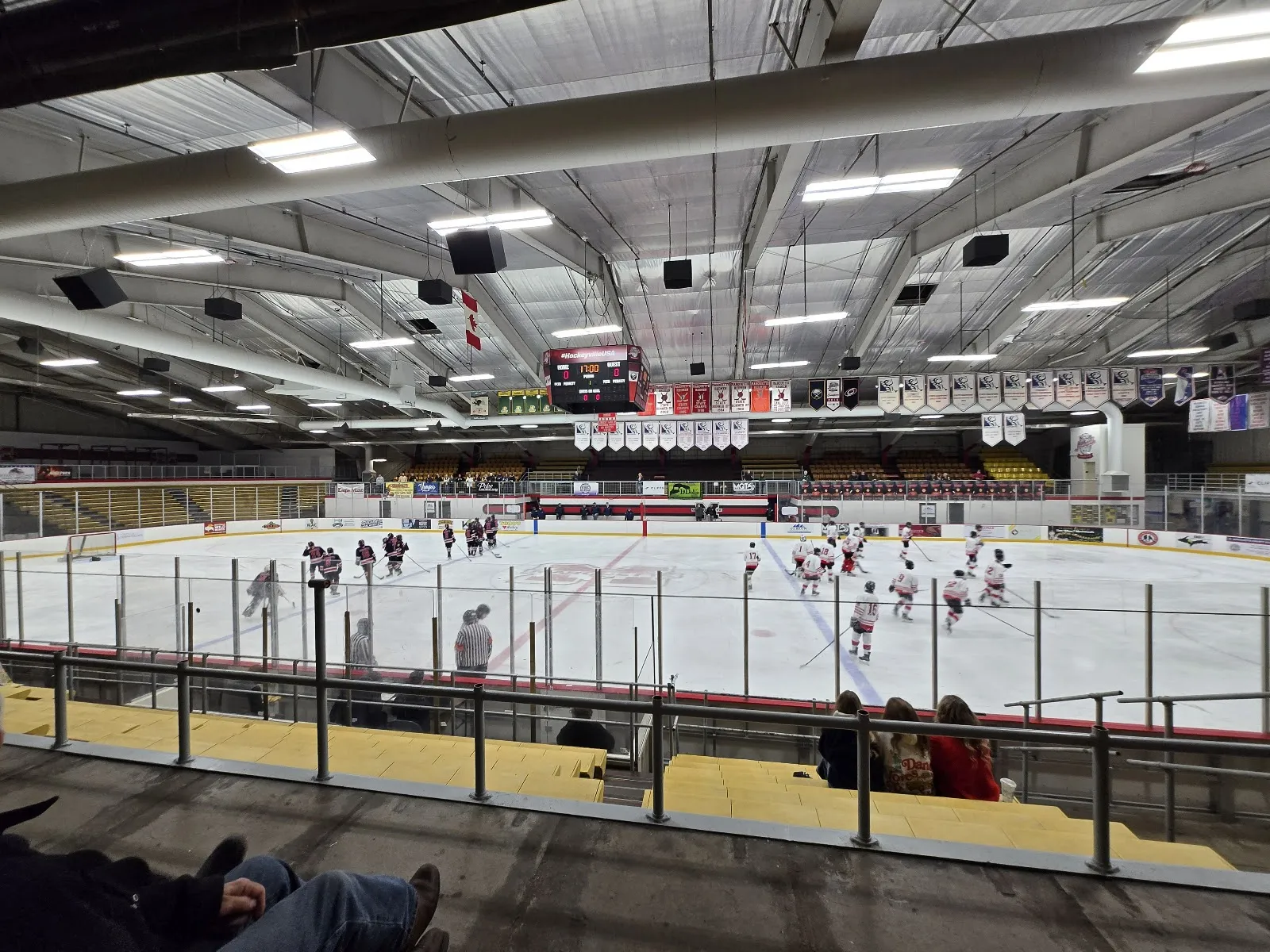 The arena bowl seen from the stands, players spread across the ice under a steel-framed roof with banners hung along the far wall and rows of bleachers in front. — Lakeland Arena