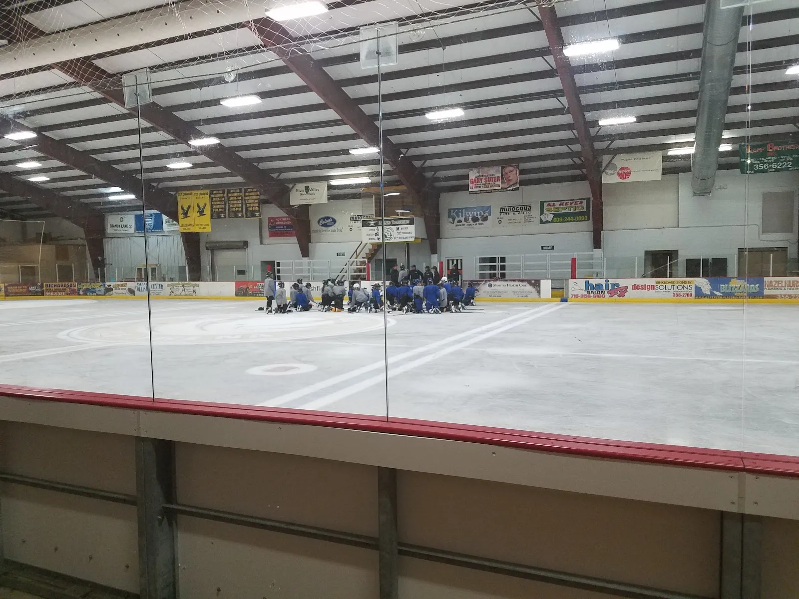 Ice sheet at Lakeland Hawks Ice Arena in Minocqua, a team gathered at the far end under the steel roof trusses — Lakeland Hawks Ice Arena