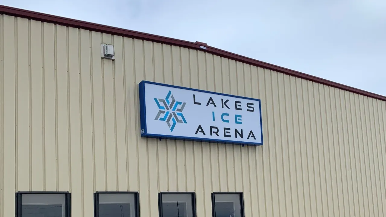 Close view of the Lakes Ice Arena building exterior, cream metal siding with a dark red roofline, a lit blue-bordered "LAKES ICE ARENA" sign with a snowflake logo mounted on the wall above ground-level windows. — Lakes Ice Arena