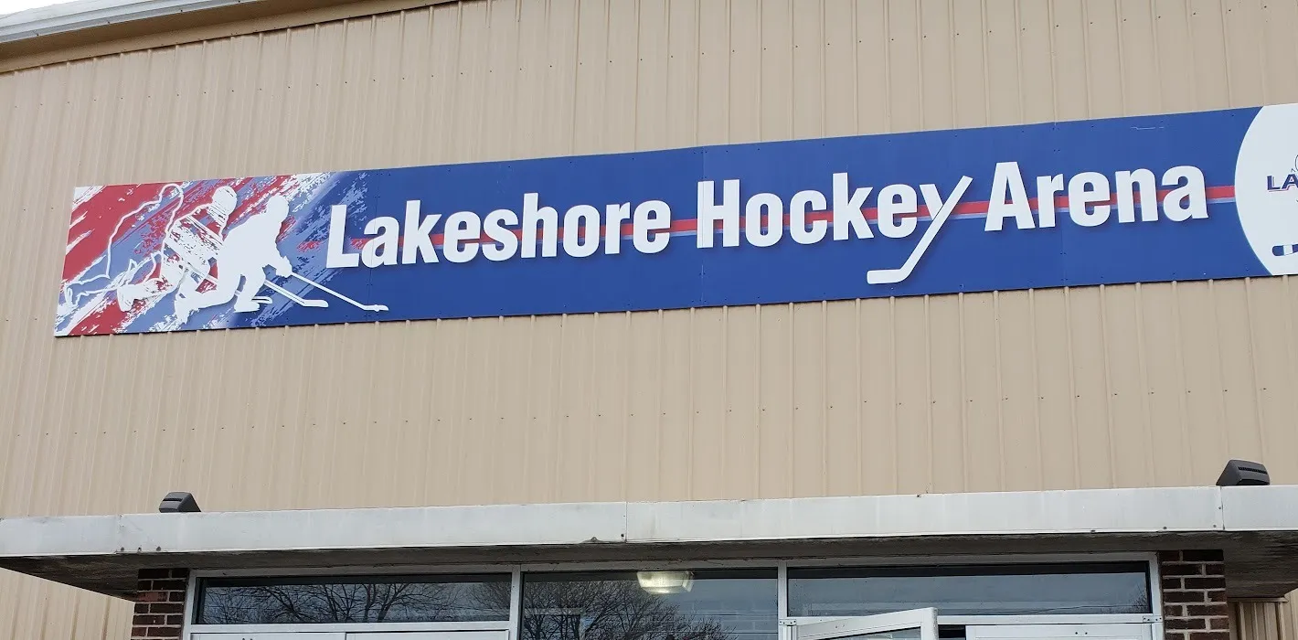 Tan metal-sided facade of the arena with a long blue banner reading "Lakeshore Hockey Arena" and a crossed-sticks Lakeshore logo, above a flat entrance canopy and the tops of the glass entry doors. — Lakeshore Hockey Arena
