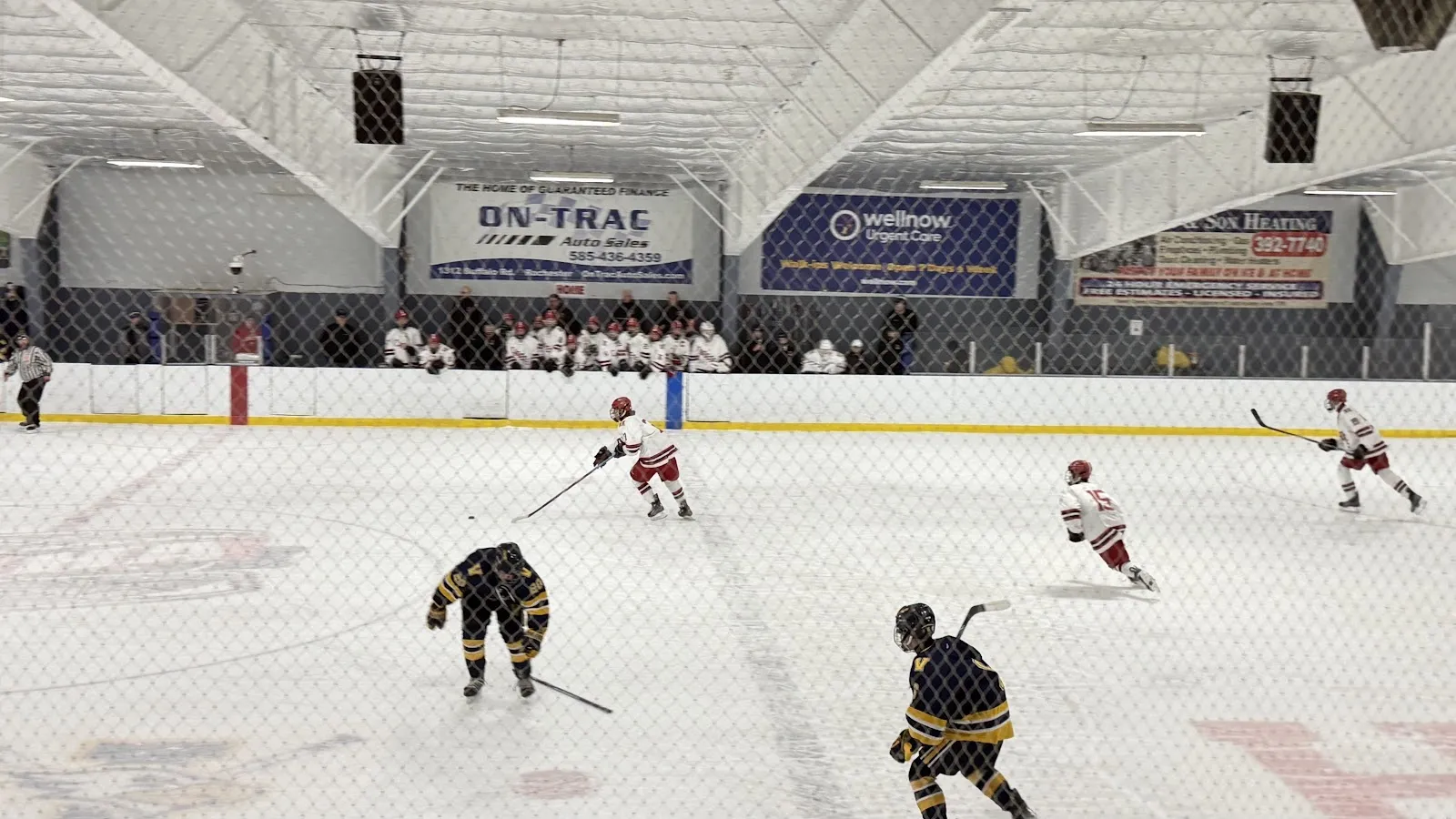 A game seen from the near glass, players spread across a wide sheet beneath a pale roof with sponsor boards along the far wall. — Lakeshore Hockey Arena