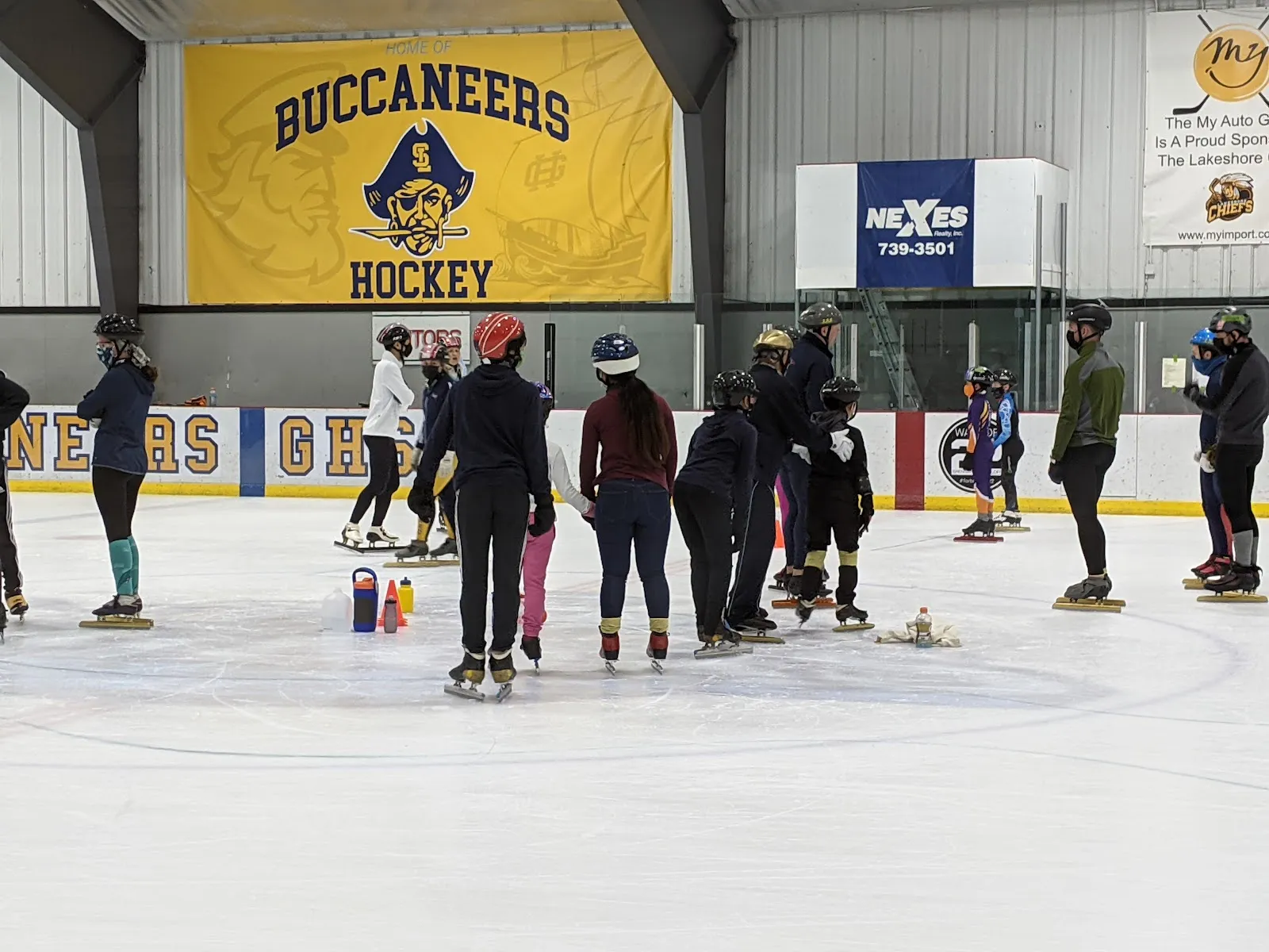 Un grupo de patinadores reunidos alrededor de conos en el centro del hielo bajo un banderín amarillo de Buccaneers Hockey, con anuncios de patrocinadores y una pared del fondo iluminada de azul detrás. — Lakeshore Sports Centre