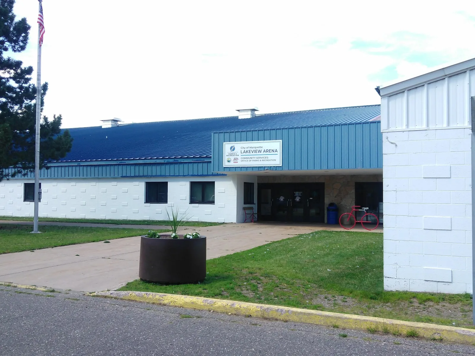 Front entrance of the building with a 'City of Marquette LAKEVIEW ARENA / Community Services: Office of Parks & Recreation' sign mounted on the blue metal facade, flagpole and bike rack out front. — Lakeview Arena