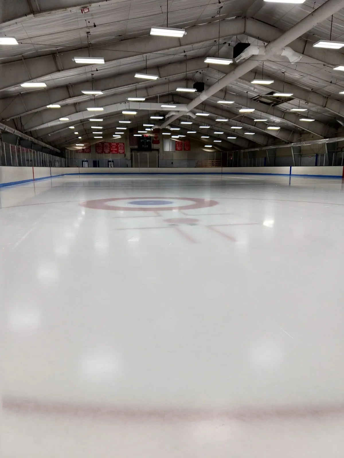 Empty full ice sheet viewed from ice level toward the far boards, with US and Canadian flags, a scoreboard and championship banners hanging from the roof trusses. — Lakeview Arena