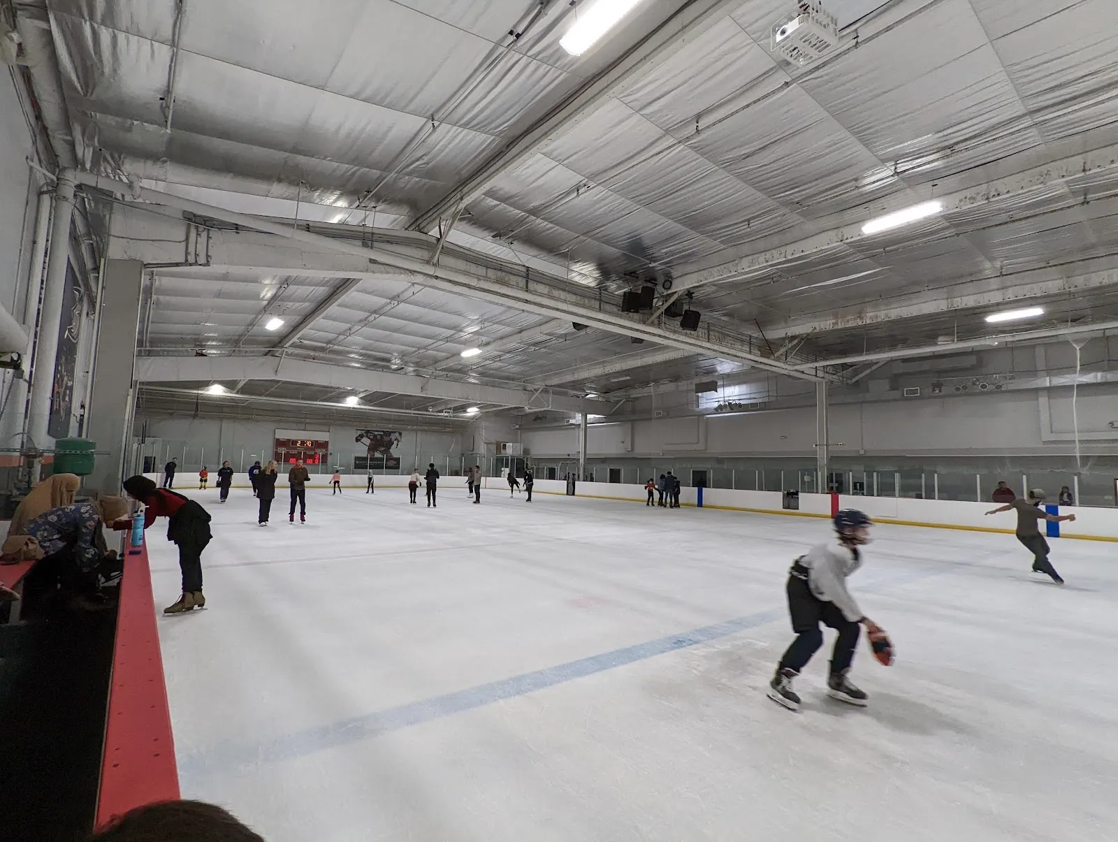 Una vista amplia de la pista con patinadores y jugadores repartidos por el hielo bajo un techo de armaduras de acero, con una valla acolchada roja en primer plano a la izquierda. — Lakewood ICE