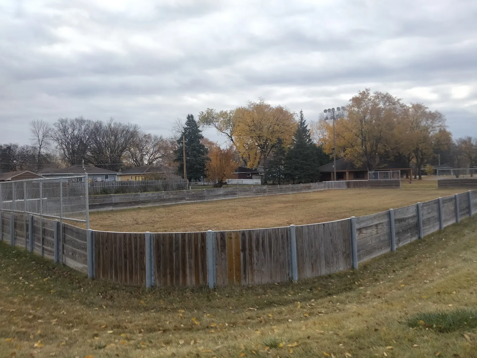 The board-enclosed rink at Lamb Park in autumn before flooding, with houses and trees beyond — Lamb Park Ice Rink