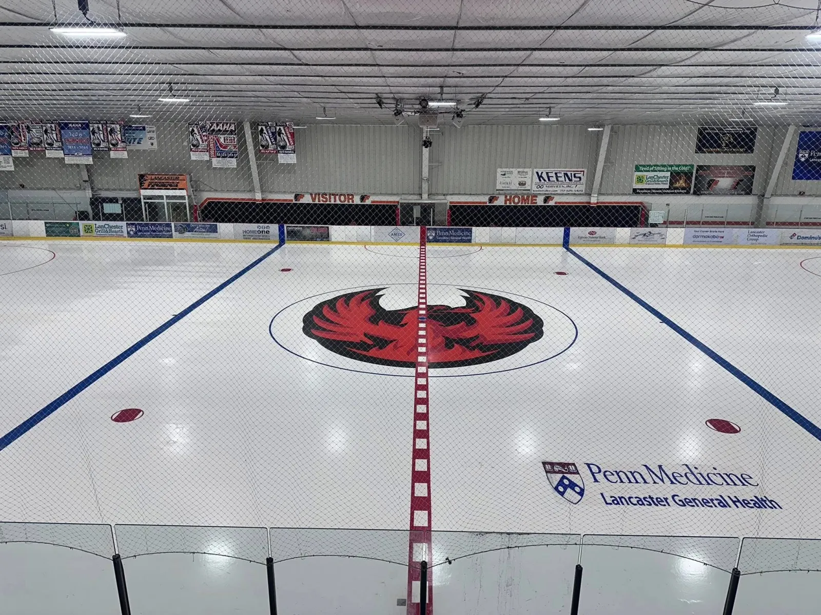 El hielo vacío visto desde un extremo, con un logotipo de un fénix rojo en el centro de la pista, un logotipo de Penn Medicine Lancaster General Health y mantas en la pared del fondo. — Lancaster Ice Rink