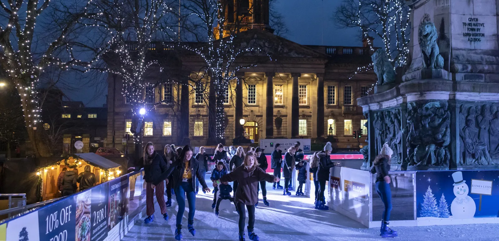Dalton Square in Lancaster at dusk with the rink in front of the town hall: skaters on the lit ice behind the boards, strings of lights in the trees and the memorial column on the right. — Lancaster On Ice (Dalton Square)