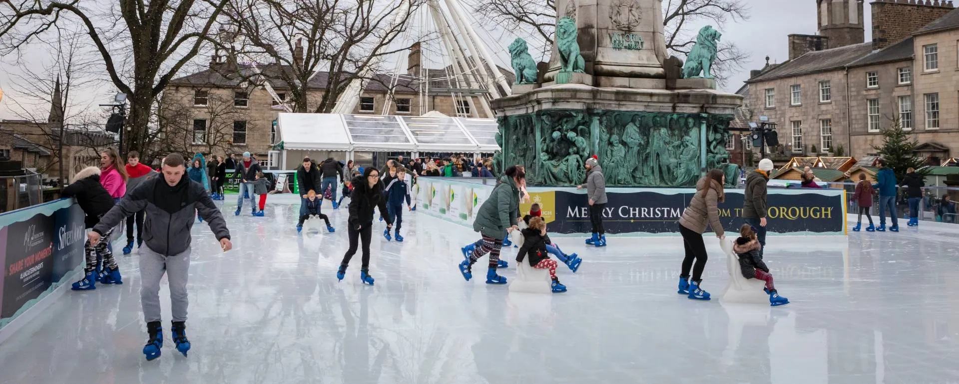 A crowd of skaters on the white ice of the open-air Lancaster rink by day, with the big wheel, the memorial statues and the stone terraces of Dalton Square behind. — Lancaster On Ice (Dalton Square)