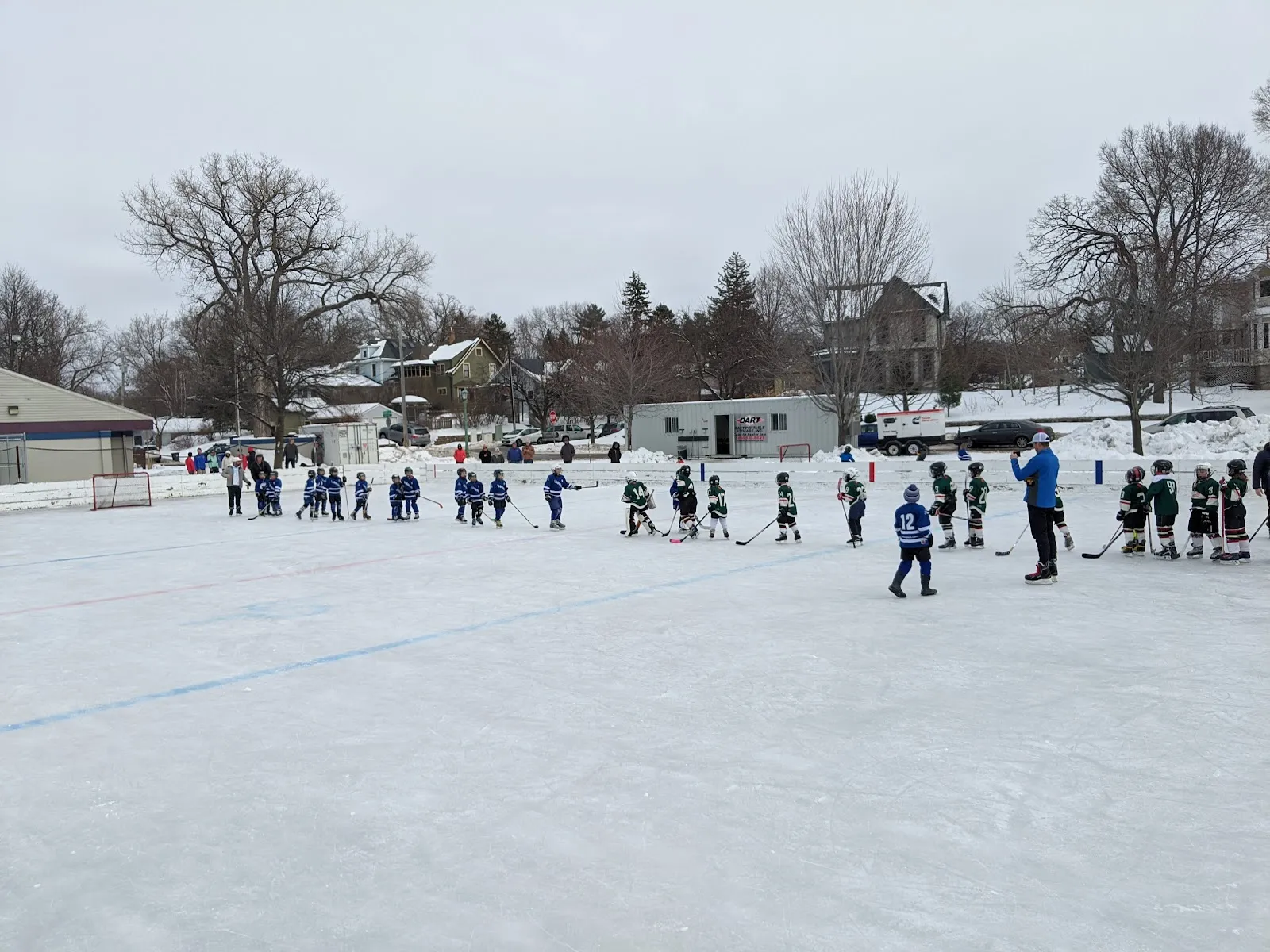 A youth hockey session on the outdoor ice at Langford Park, with players spread along the far side, snowbanks and bare trees behind. — Langford Park Outdoor Rinks
