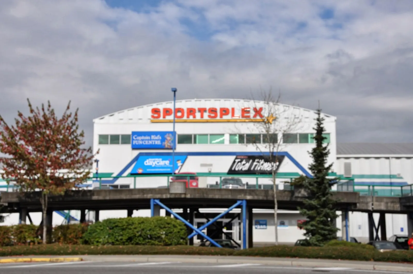 The front of the Langley Sportsplex under a cloudy sky, with the large Sportsplex sign above the daycare and fitness entrances and trees in front. — Langley Sportsplex