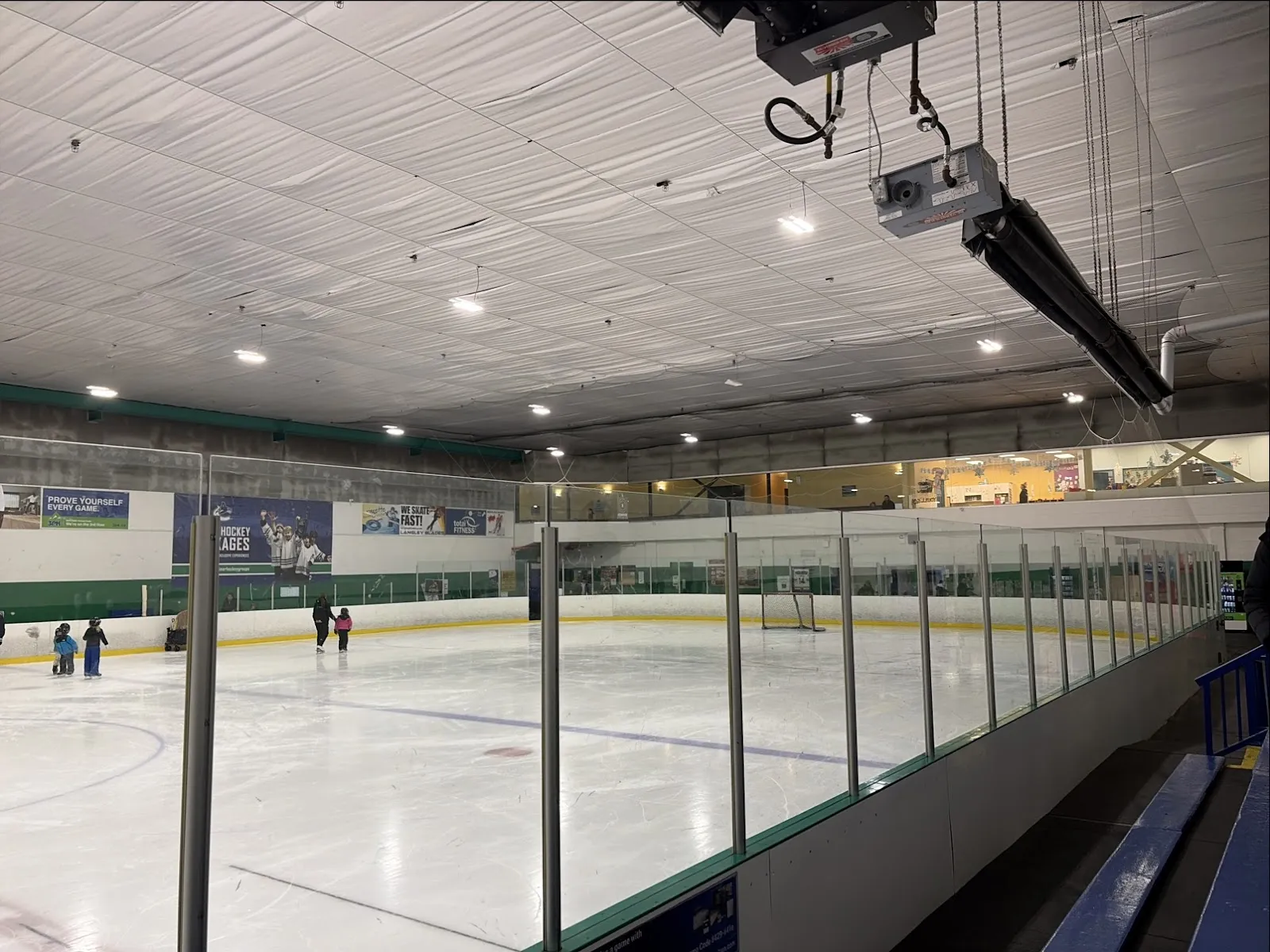 One of the Sportsplex rinks with a few skaters on the ice, the glass and netting along the boards and the lit ceiling above. — Langley Sportsplex