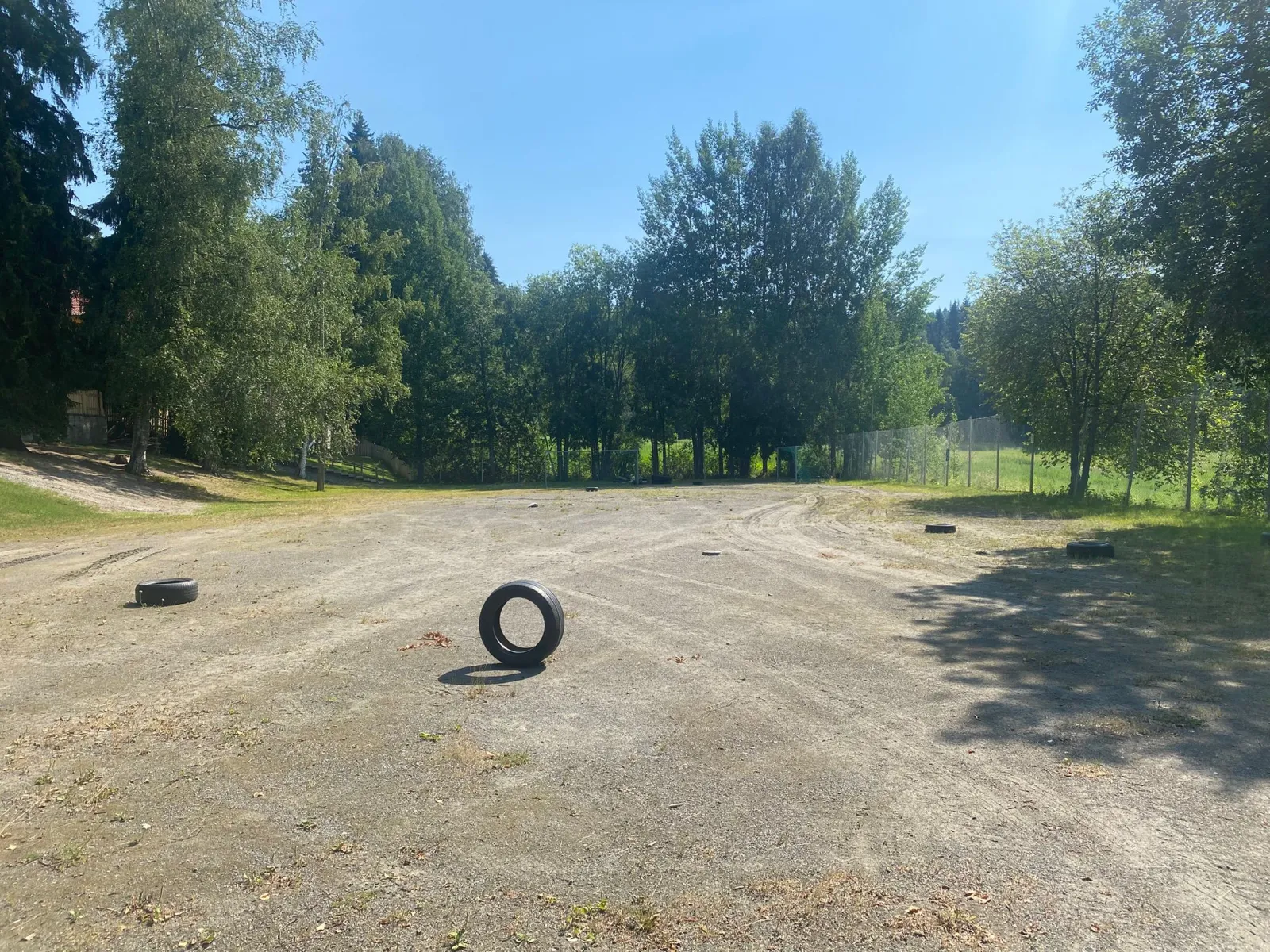 The sandy field beside the Länsi-Puijo daycare in Kuopio, which serves as a skating field in winter, seen in summer with a few old tyres on the ground and trees behind. — Länsi-Puijon päiväkodin luistelukenttä