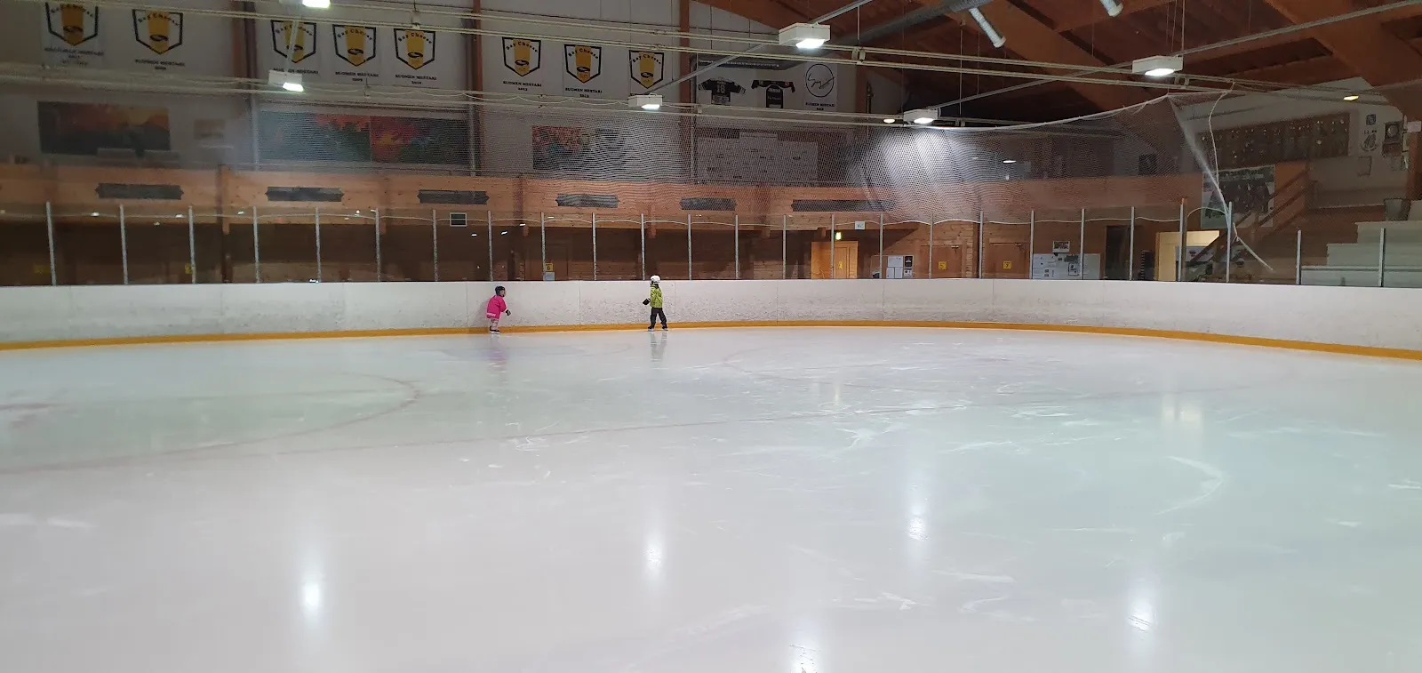 Two skaters far across the pale ice of the Lapinlahti ice hall, with the boards and tiered roof lights behind. — Lapinlahden jäähalli