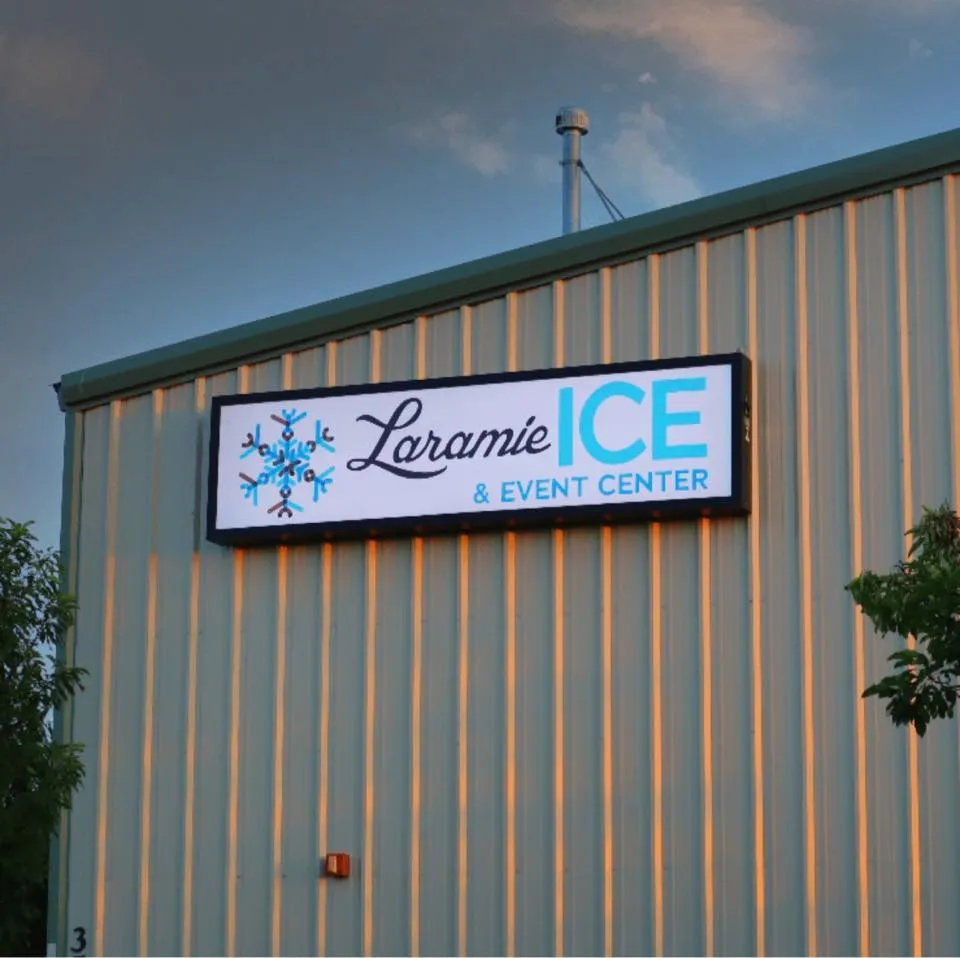 Low-angle view of the building's metal-panel gable end at dusk, with the lit 'Laramie ICE & EVENT CENTER' sign mounted on the wall below the roofline. — Laramie Ice and Events Center
