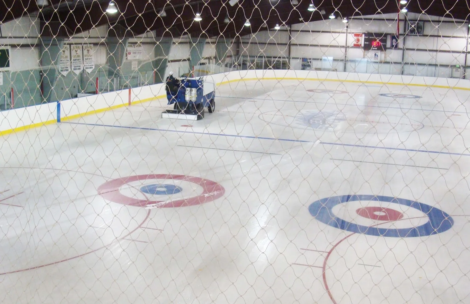 A wide view of the sheet with the centre circle in the foreground, a few players at the far boards under a steel roof with banner strips. — Laramie Ice and Events Center