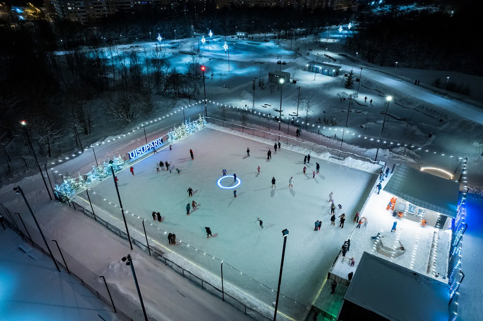 An evening view from above of the Lasnamäe refrigerated outdoor rink, a fenced rectangle of lit ice beside its low service building, with the Lasnamäe housing blocks behind. — Lasnamäe uisupark