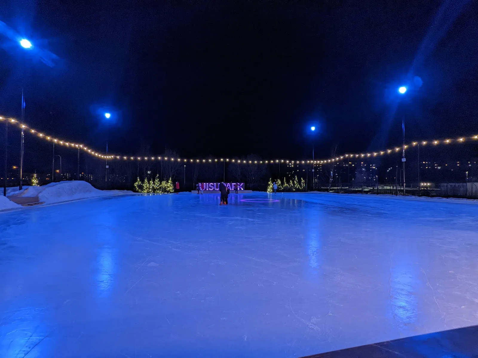 The empty ice of the Lasnamäe outdoor rink after dark, lit by pole lamps and a string of lights along the fence. — Lasnamäe uisupark