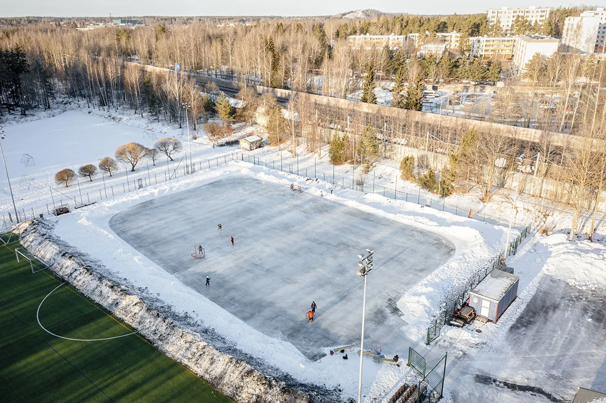 Flygbild över naturisbanan i Lassila idrottspark, med några skridskoåkare på isen bredvid en fotbollsplan och en snötäckt skog. – Lassilan liikuntapuisto – luonnonjää