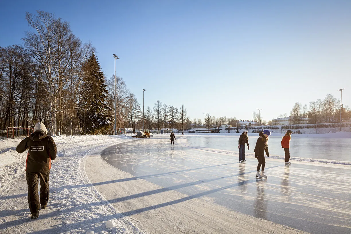 A curve of the skating rink in Lauttasaari sports park on a clear winter day, with a few skaters on the ice and bare birches along the edge. — Lauttasaaren liikuntapuisto – luistelukenttä