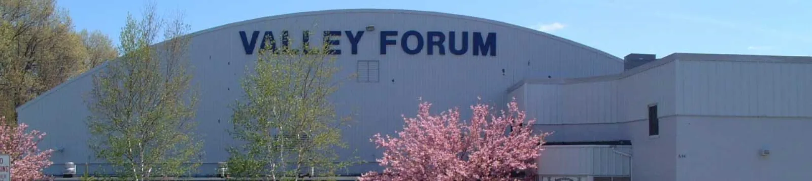 Edificio de la arena con revestimiento metálico blanco y techo en arco, las letras "VALLEY FORUM" en la fachada y árboles en flor al frente, fotografiado en un día despejado — Lawrence Valley Forum