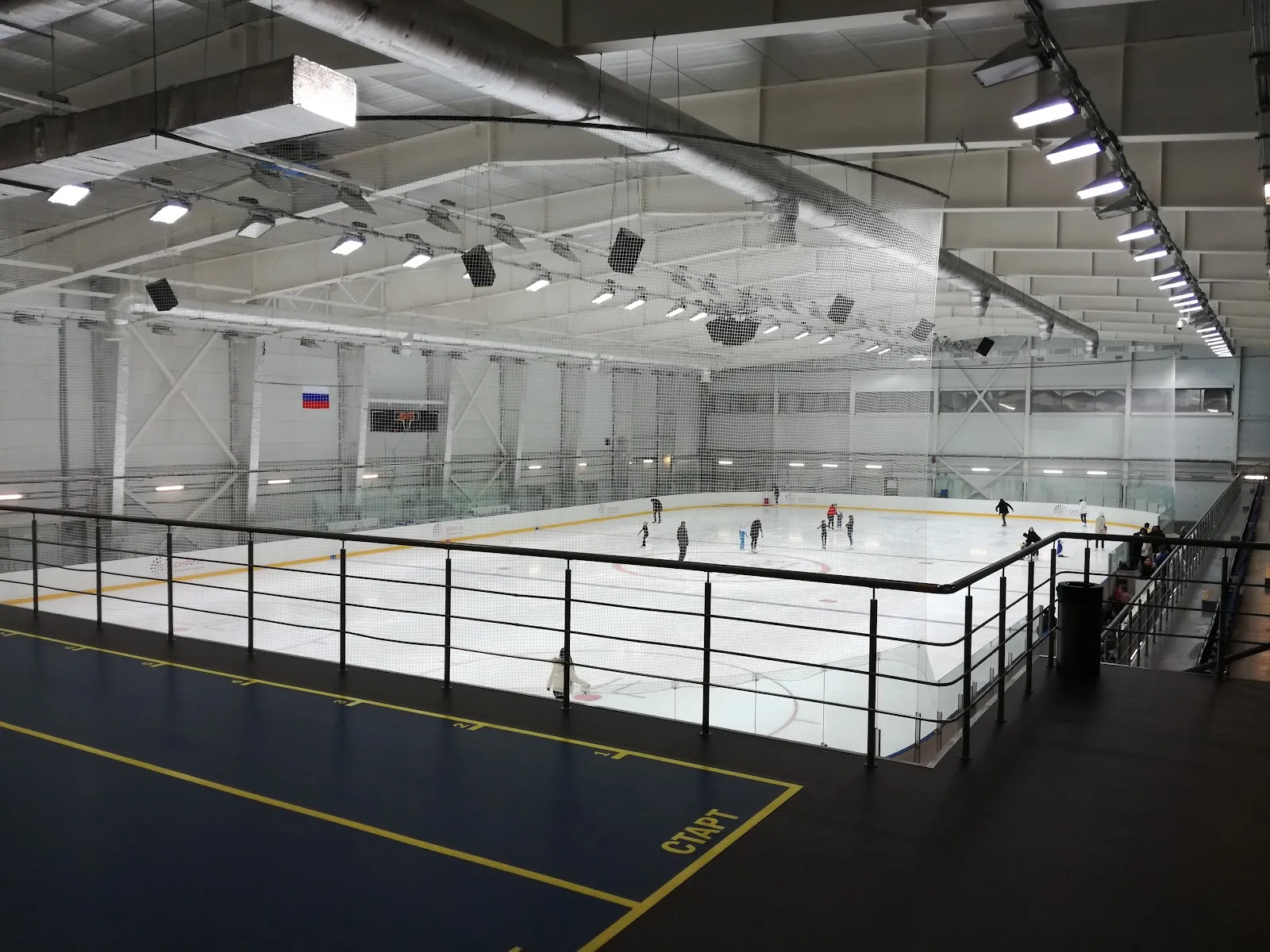 The ice sheet of the Lyodinka training arena in Sirius seen from the upper gallery, with a few skaters on the ice and the roof trusses and lighting above — Lyodinka Ice Arena Sirius