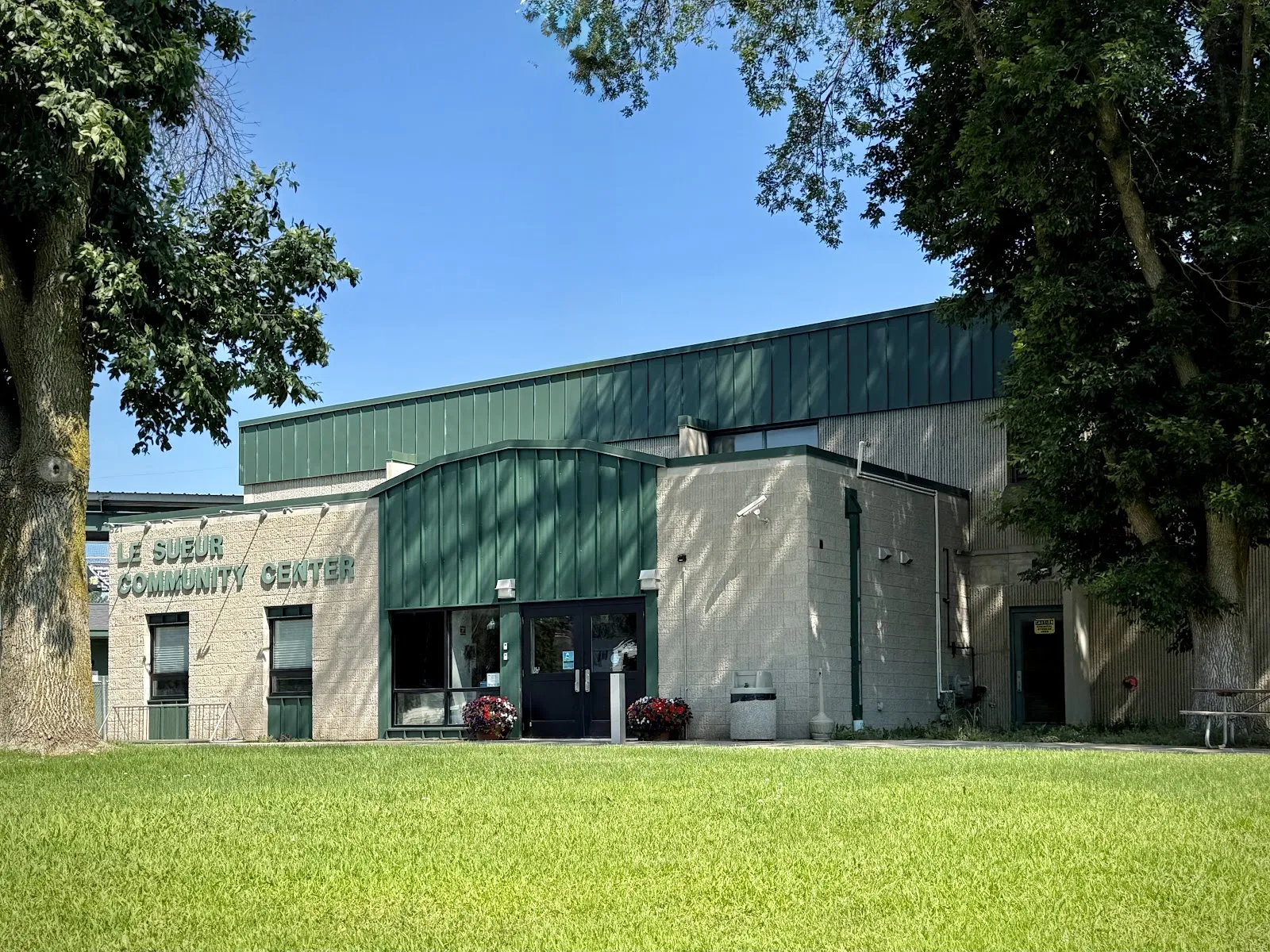 The green-roofed Le Sueur arena building seen across the grass, with trees beside it. — Le Sueur Community Center Ice Arena