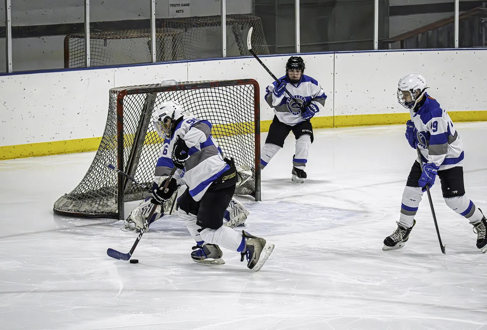 Youth hockey play in front of the net on the indoor ice at Le Sueur, with the boards and rink glass behind. — Le Sueur Community Center Ice Arena