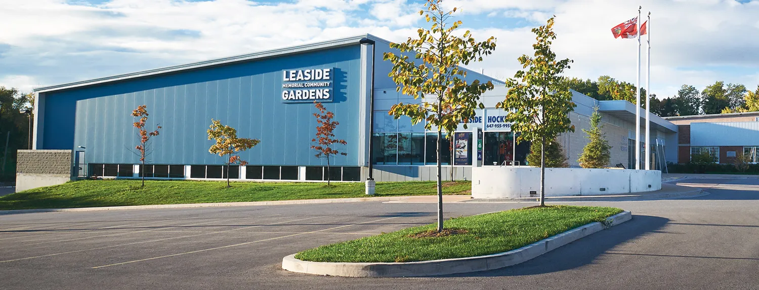 The newer blue-clad rink building with the "Leaside Memorial Community Gardens" sign, young trees and a flagpole in front. — Leaside Memorial Community Gardens
