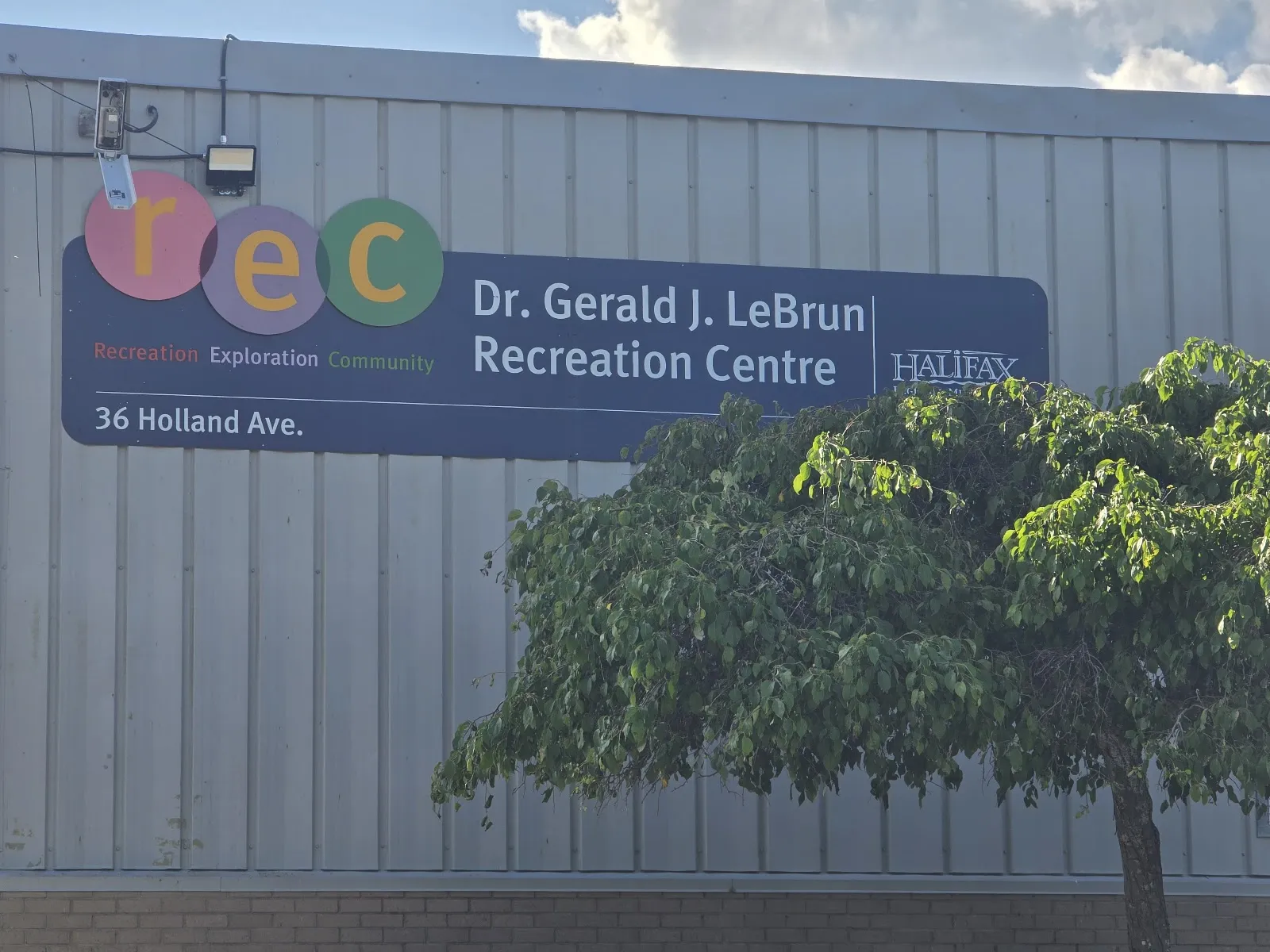 The Dr. Gerald J. LeBrun Recreation Centre sign with its 36 Holland Ave address on the grey metal wall of the building, with a tree in front. — LeBrun Arena