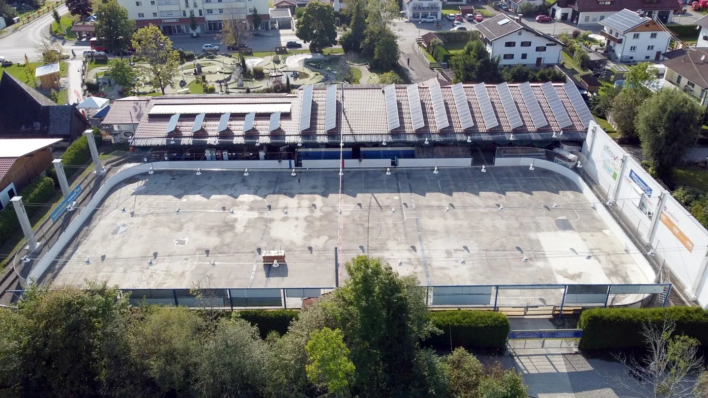 The Lechparkstadion in summer, seen from the air: the bare concrete rink floor, the grandstand with its sawtooth roof and the village behind. — Lechparkstadion Lechbruck