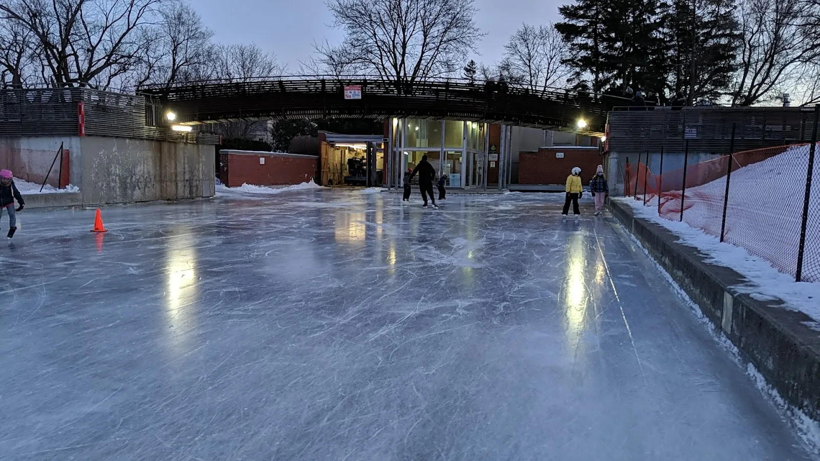 Ledbury Park rink ice at dusk with the boards, ramp walkway and pedestrian bridge behind — Ledbury Park Outdoor Ice Rink