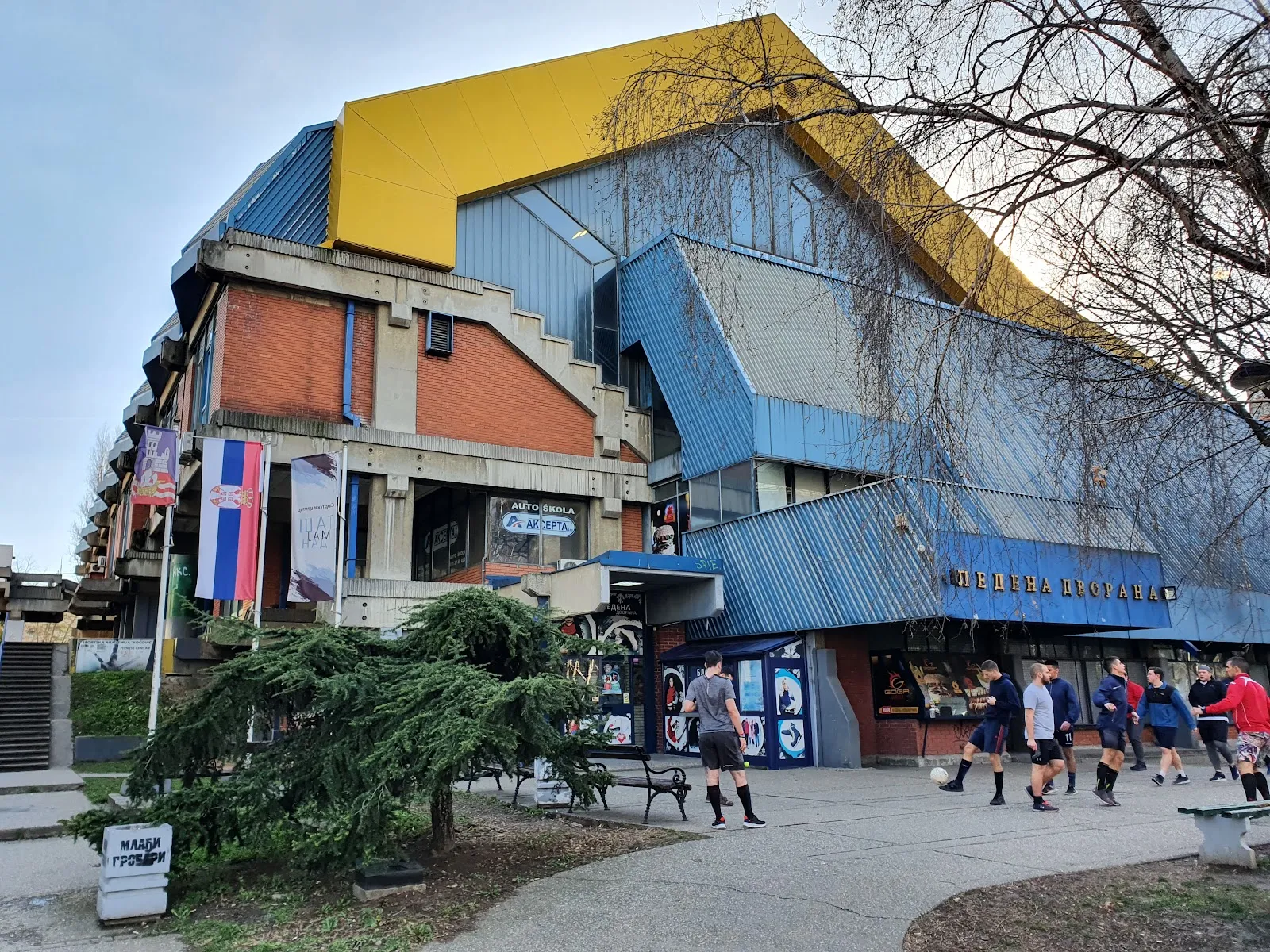 The blue-clad ice hall with its yellow roof edge and the entrance plaza in front, seen from the park path — Ledena dvorana Pionir