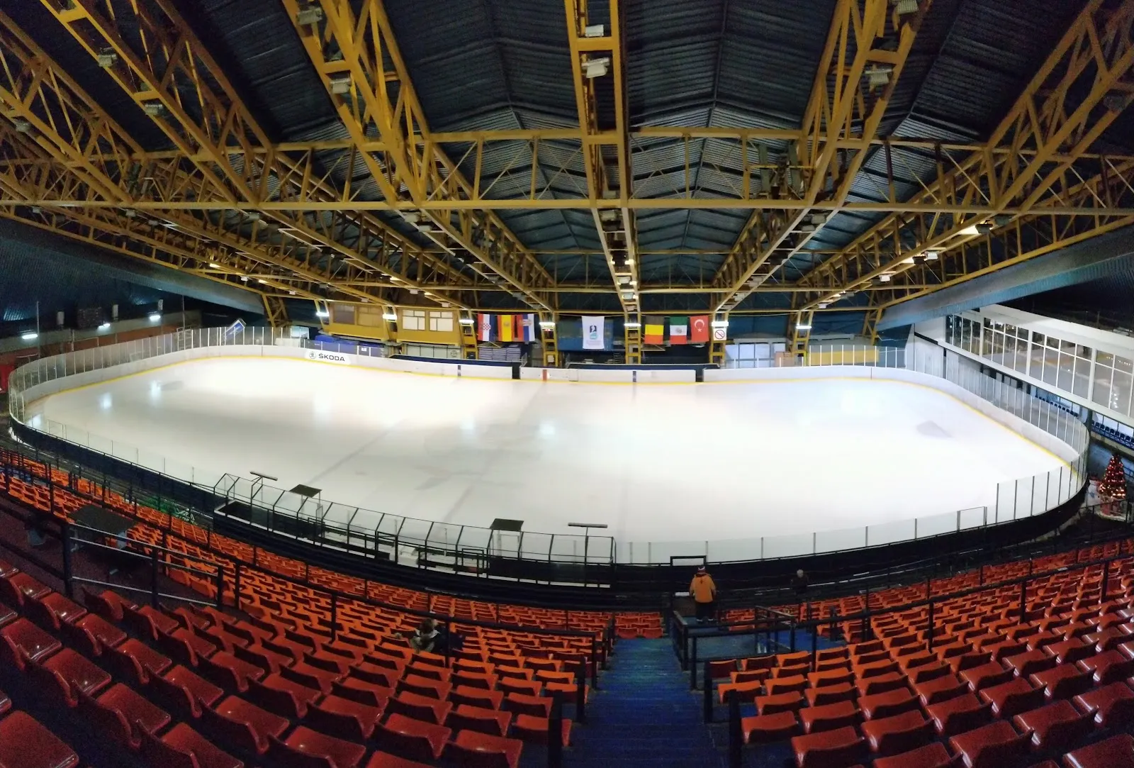The empty ice sheet seen from the stands, under the yellow steel roof trusses, with rows of orange seats along the boards — Ledena dvorana Pionir