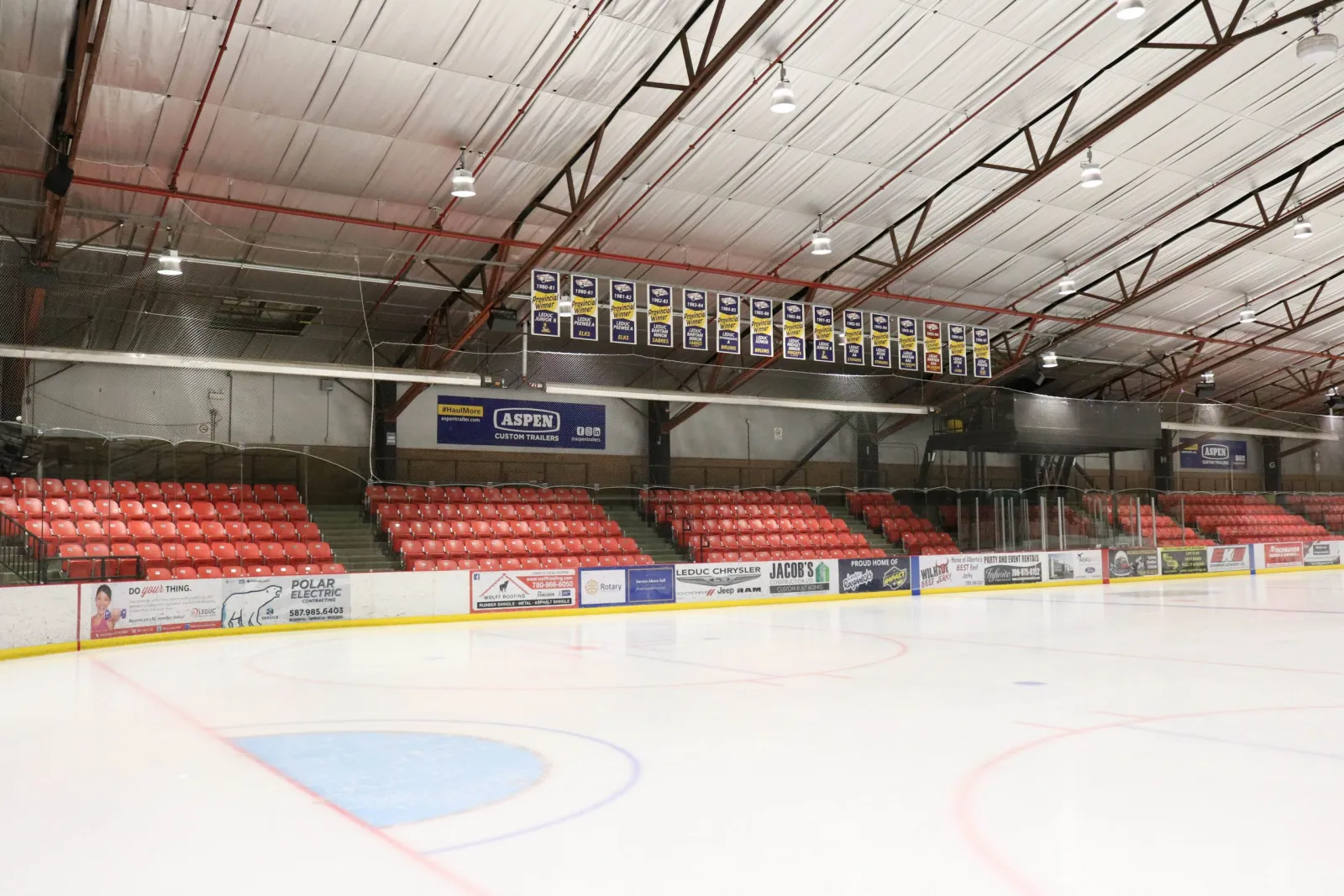 The empty Aspen Custom Trailers Arena under its steel roof, with red seats along the side and championship banners overhead. — Leduc Recreation Centre
