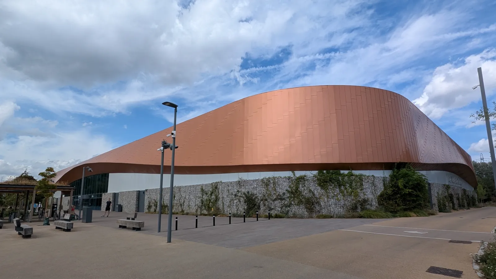 The rebuilt Lee Valley Ice Centre on Lea Bridge Road, its curved copper-coloured roof above a planted stone wall and the entrance plaza. — Lee Valley Ice Centre