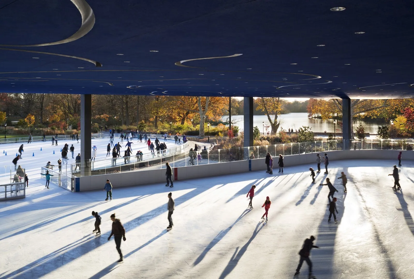 Patinadores repartidos por la pista techada bajo un toldo azul oscuro, con árboles otoñales y el lago más allá de los costados abiertos. — LeFrak Center at Lakeside