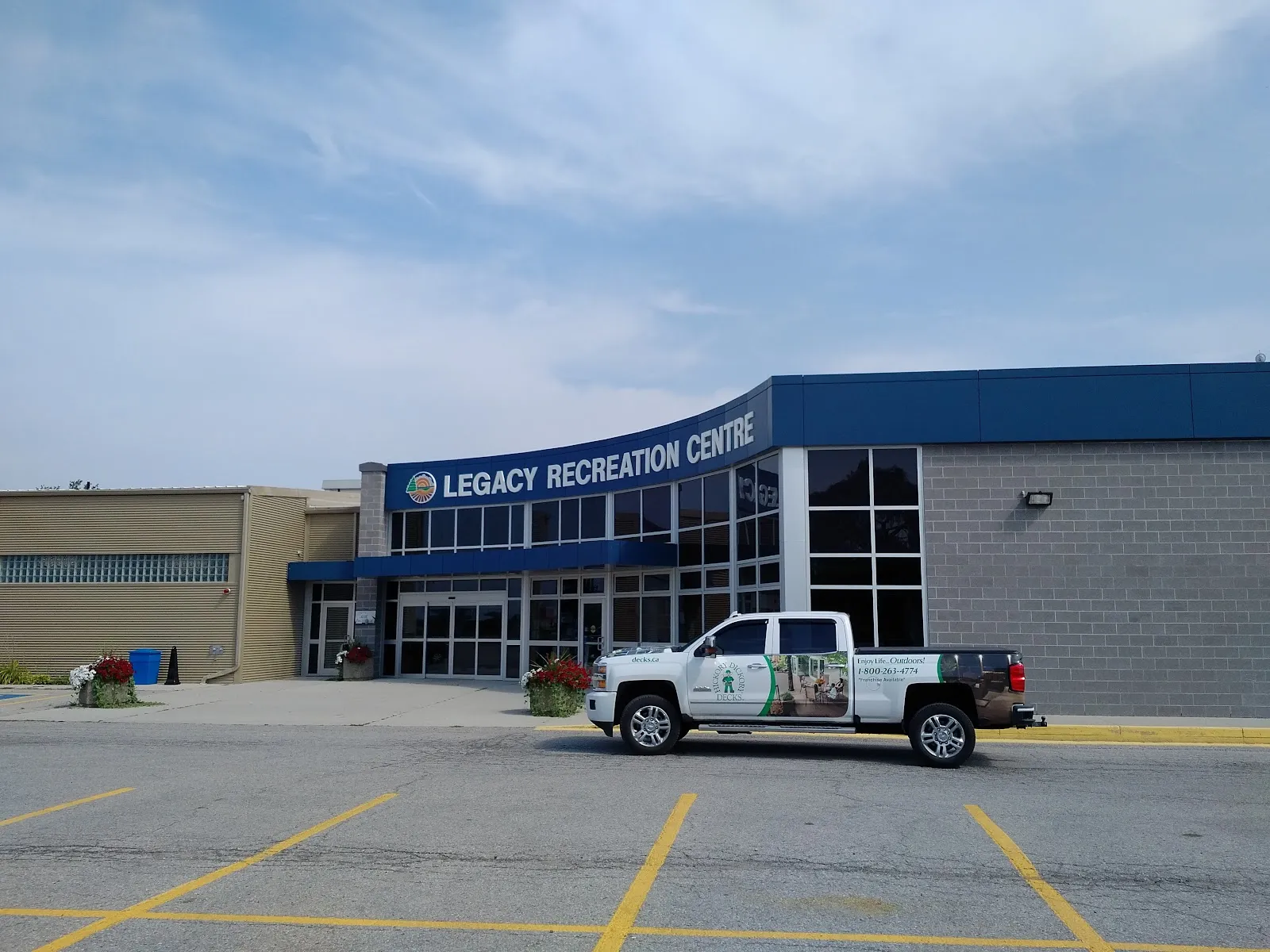 La entrada de vidrio del Legacy Recreation Centre bajo un letrero azul, con sus muros de bloque gris junto al estacionamiento. — Legacy Recreation Centre (Thedford)