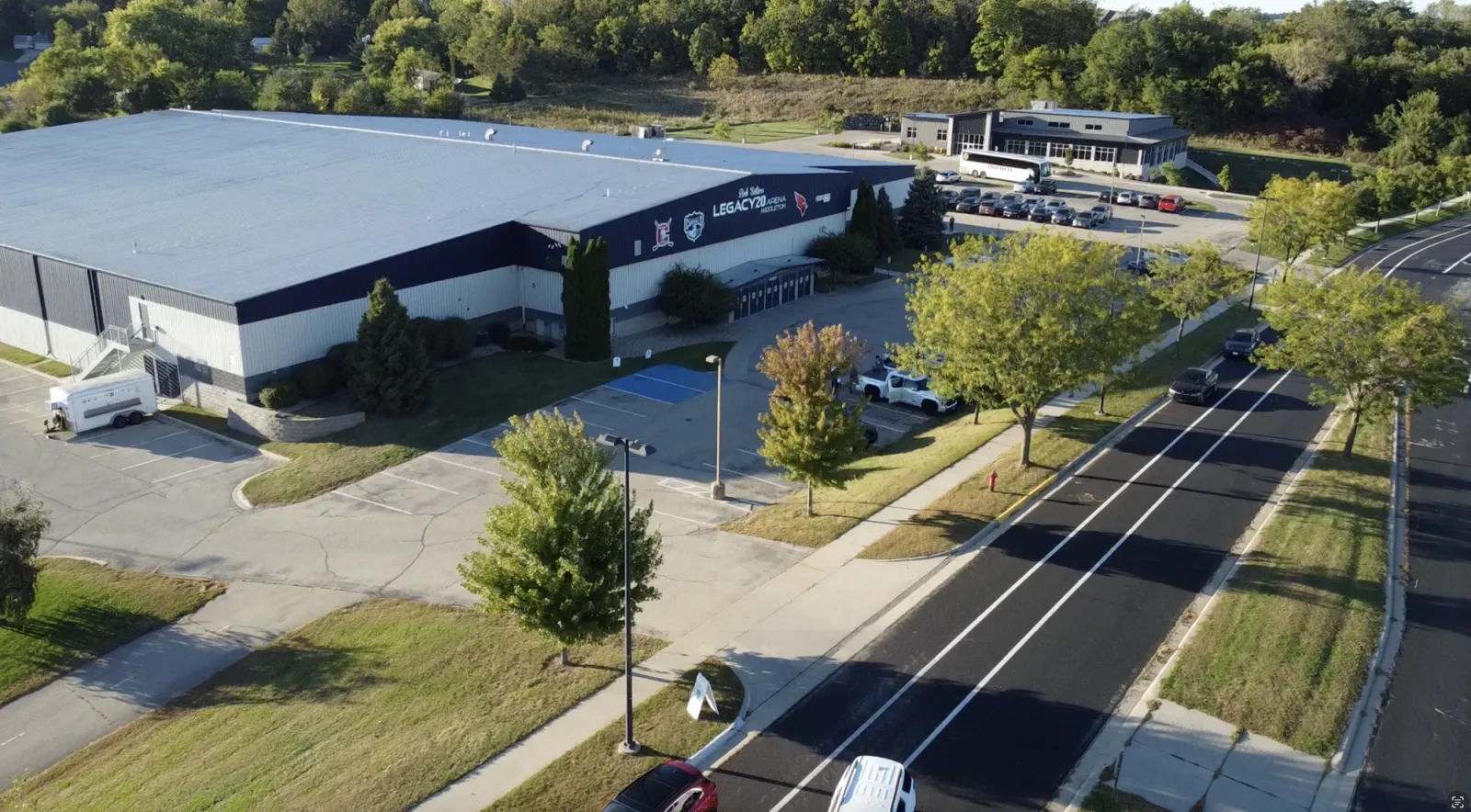 Aerial view of LEGACY20 Arena Middleton, its dark blue and white metal-panel building with the arena name and Cardinal/Capitols logos painted on the gable end, parking lot and entrance canopy in front. — LEGACY20 Arena Middleton