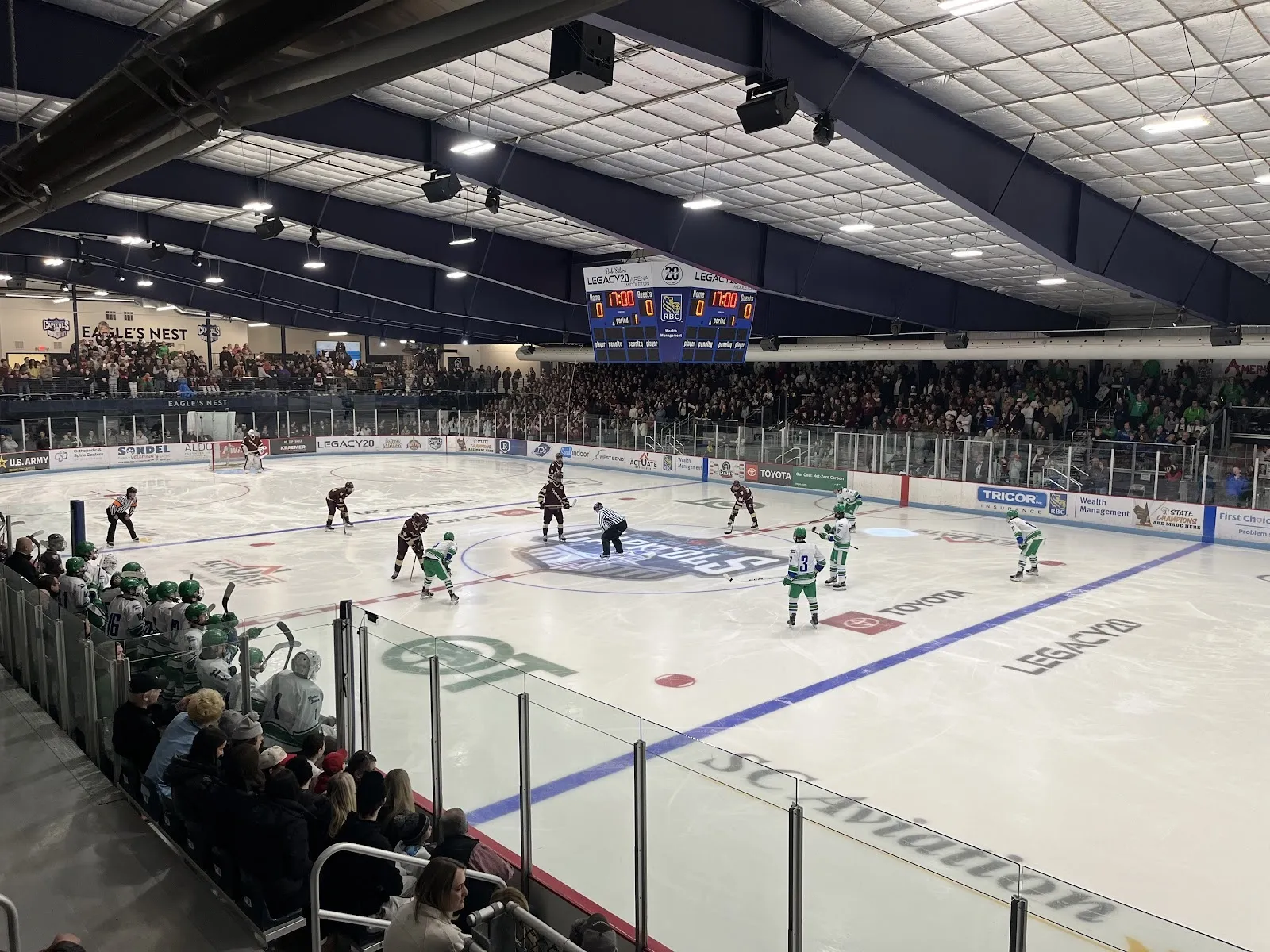 A faceoff in front of the stands, players spread across the ice with a crowd packed along the glass beneath a dark blue-trussed roof. — LEGACY20 Arena Middleton