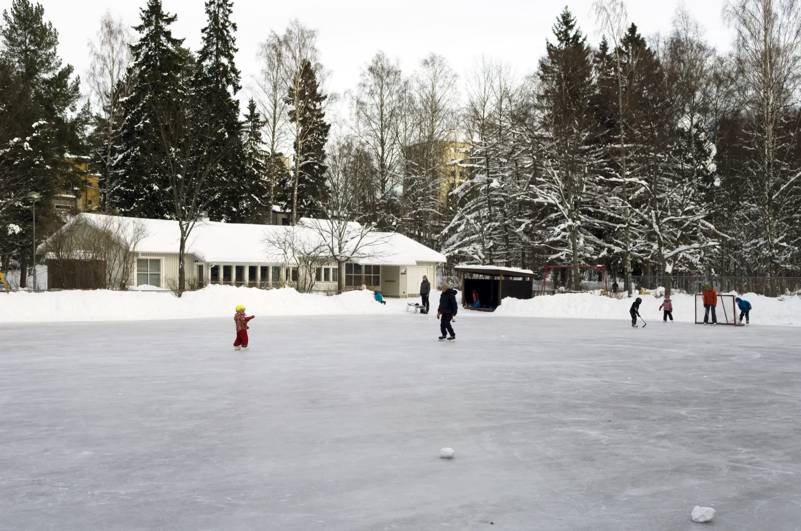 Ulvilan leikkipuiston luistelukenttä Munkkivuoressa Helsingissä tammikuussa: jäällä lapsia ja muutamia luistelijoita, taustalla matala valkoinen rakennus ja lumisia kuusia. – Leikkipuisto Ulvila – luistelukenttä