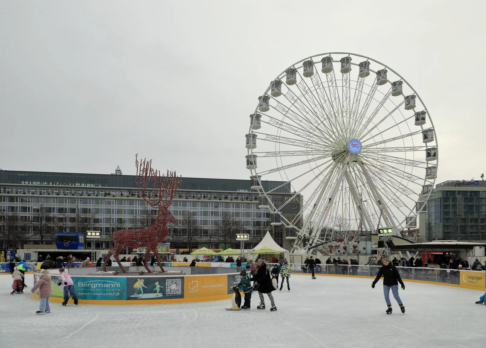 Skaters of all ages on the round ice of the Leipziger Eistraum in February 2026, with the lit-up Ferris wheel and a large antler sculpture on the Augustusplatz. — Leipziger Eistraum