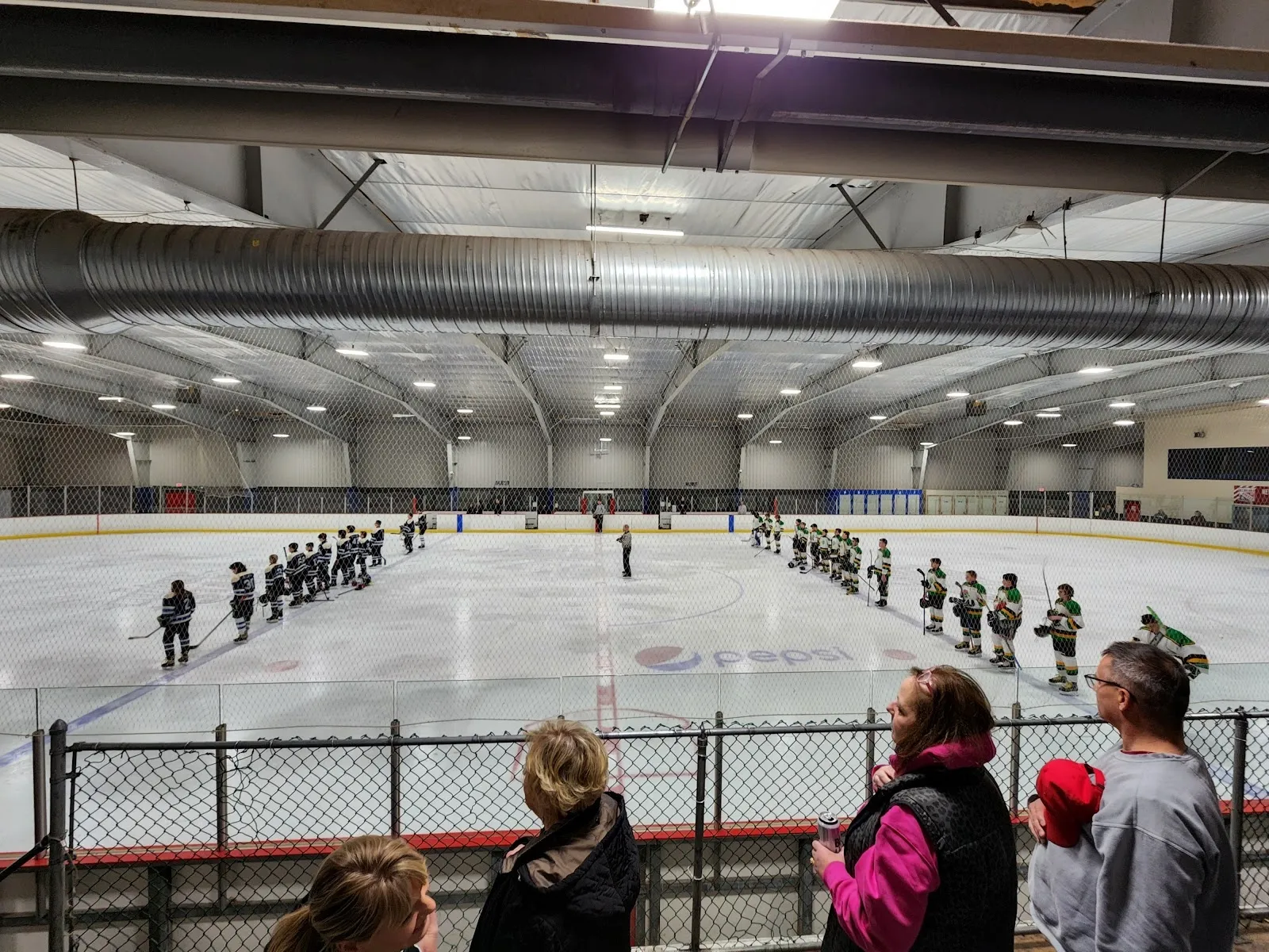Players lined up on the blue lines across a wide sheet before a game, parents watching at the rail under a dark-trussed ceiling. — Leisure Rinks