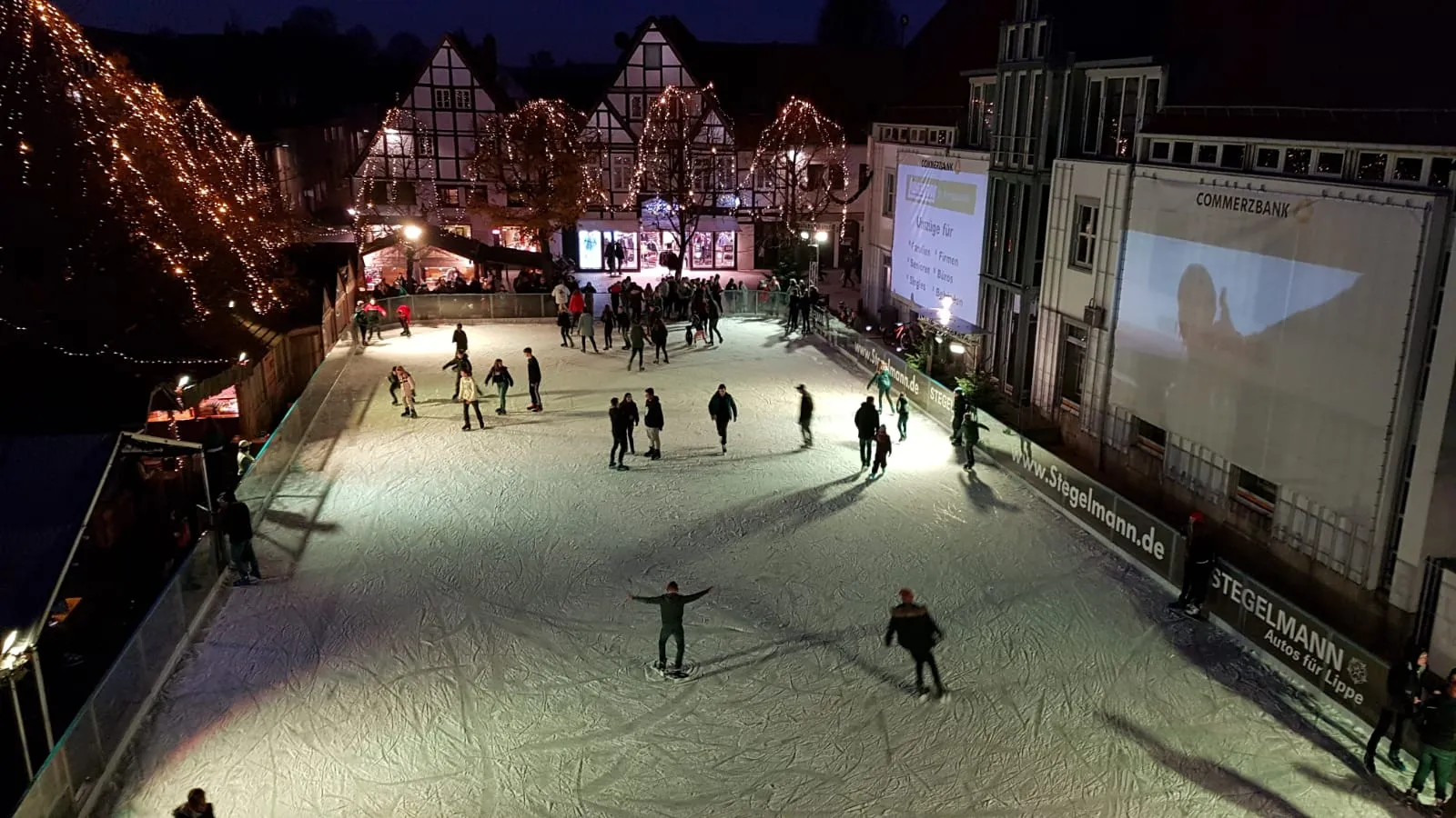 Luftaufnahme der Lemgoer Eiswelt im Lippegarten bei Nacht: kleine Schlittschuhläufer auf dem beleuchteten Eis, ringsum Fachwerkhäuser und festliche Beleuchtung. – Lemgoer Eiswelt