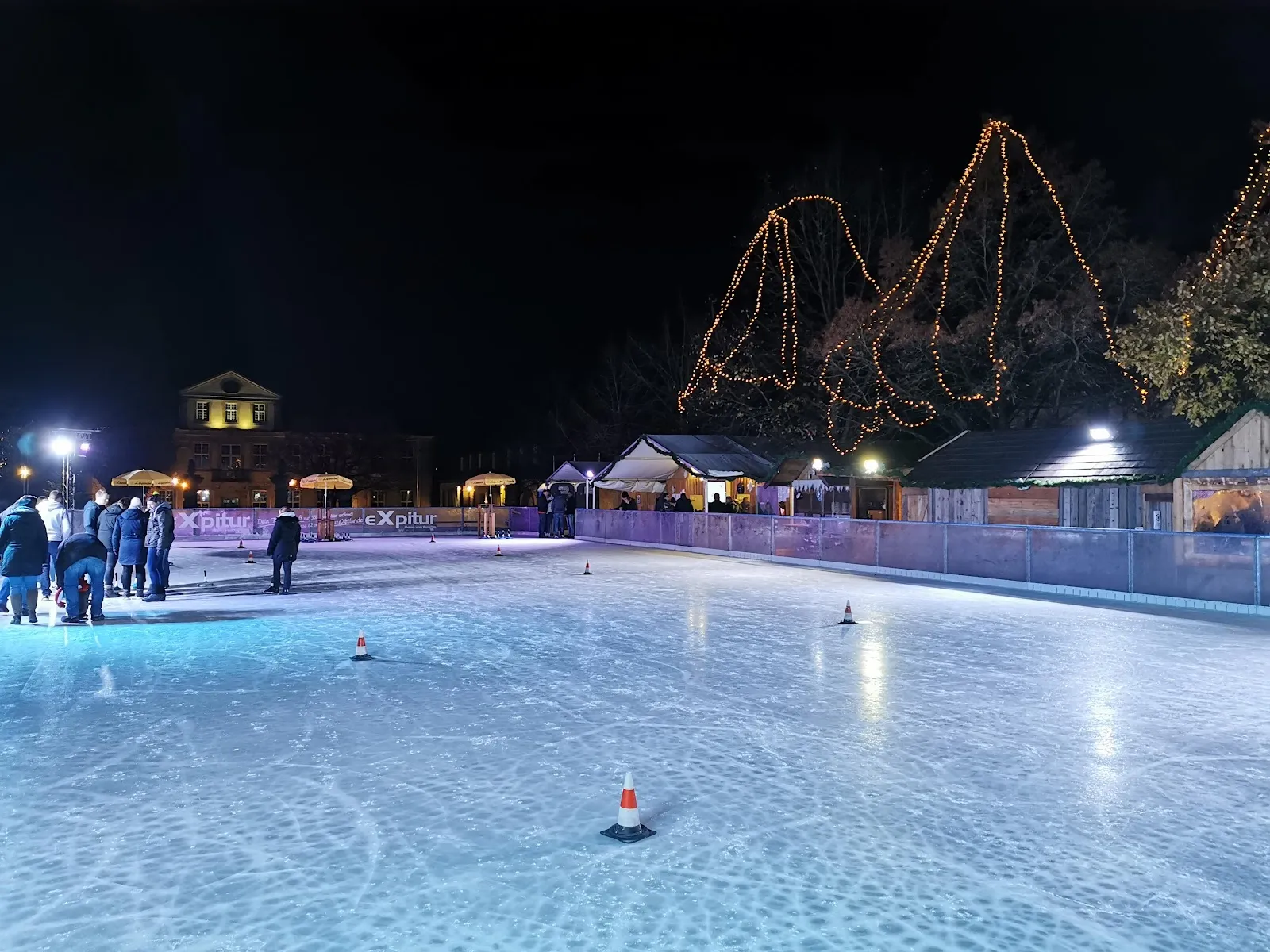Das Freiluft-Eis der Lemgoer Eiswelt bei Nacht, von Flutlicht beleuchtet, mit einigen Schlittschuhläufern am anderen Ende, Banden und Lichterketten am Rand und den Markthütten dahinter. – Lemgoer Eiswelt