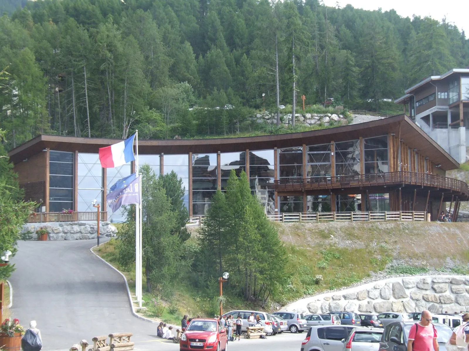The Les Orres rink building with its long timber-clad roof and a French flag on a pole, seen from the car park among pine trees. — Patinoire des Orres