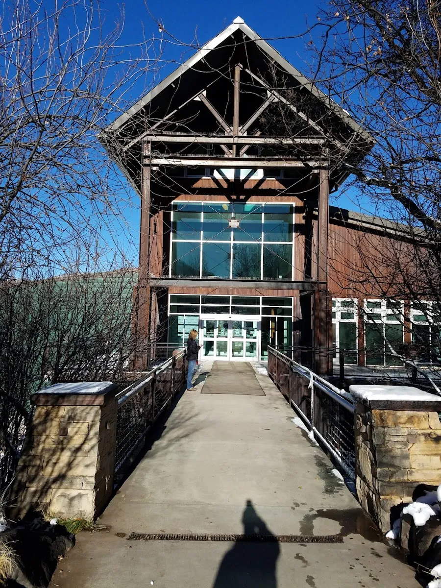 The timber-framed, glass-fronted entrance of the Aspen Recreation Center, reached by a footbridge with stone pillars. — Lewis Ice Arena