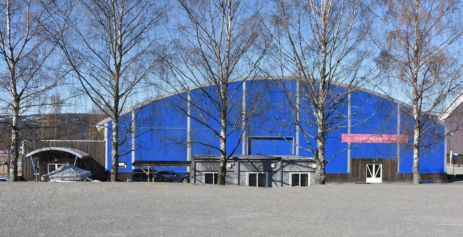 The blue arched front of Lillehammer ungdomshall seen through bare birch trees, with its timber-clad entrance annex at the foot of the hall. — Lillehammer ungdomshall