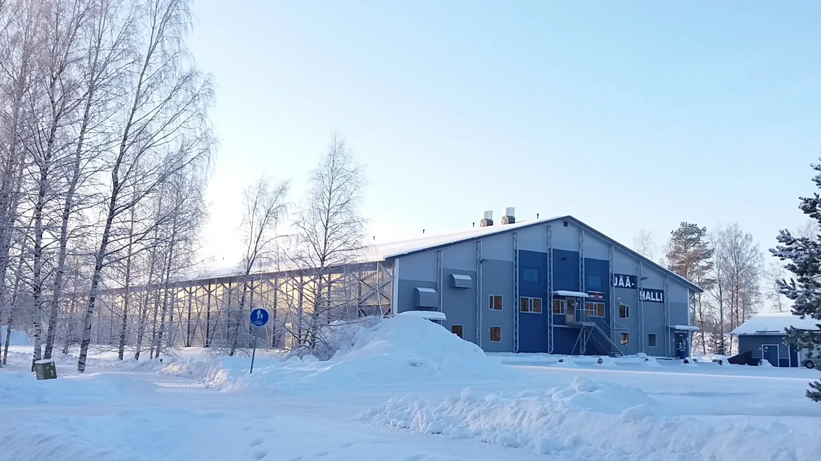 The Liminka ice hall in winter, a blue-grey building beside snow-covered birches – Limingan Jäähalli