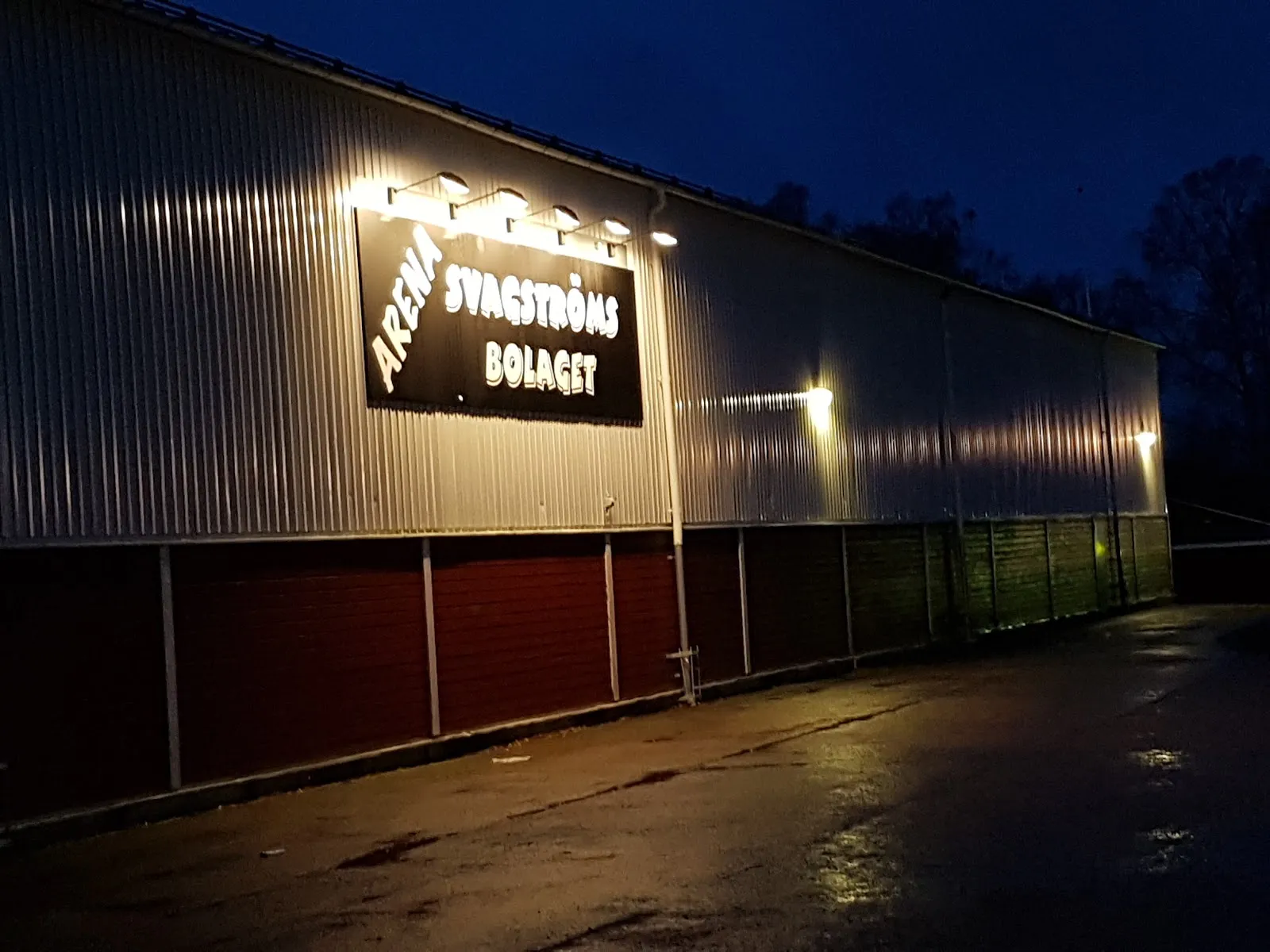 Ishallens plåtklädda sidovägg upplyst på natten, med skylten från ett tidigare sponsornamn, Arena Svagströms Bolaget – Lindbergs arena